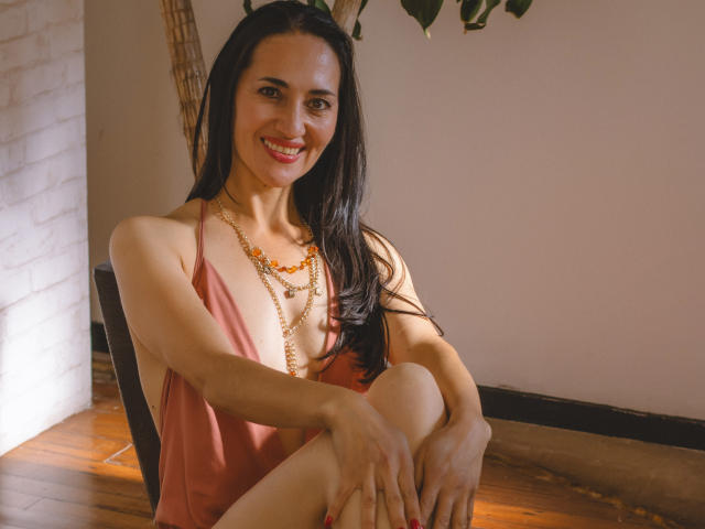 MiaMott met lang zwart haar, lachend, gekleed in een roze jurk, met een gouden ketting en oranje kralen, zittend met de knieën opgetrokken, houten vloer, en een plant op de achtergrond..