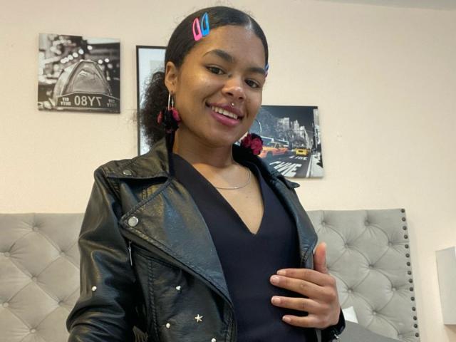 LucyLars, donna di carnagione scura con capelli ricci, mollette rosa per capelli, top nero a V, giacca di pelle, che tiene la pancia, sorridente, orecchini rosa, letto nero con testiera imbottita, selfie casual..