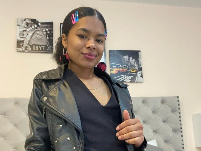 LucyLars, con piel oscura, pinzas para el pelo rosas, una camiseta negra de cuello en V, una chaqueta de cuero negra y aros de aro rosas, levantando el pulgar, sonriendo, con lápiz labial rosa, en un dormitorio..