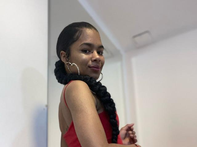LucyLars, avec sa peau foncée, ses longs cheveux noirs tressés, un débardeur rouge, montrant son dos, portant de grandes boucles d'oreilles, souriant à la caméra, regardant par-dessus son épaule..