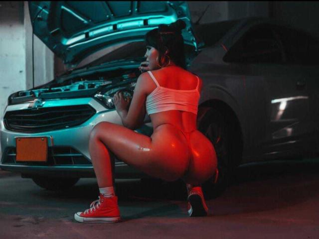 Heyly, with shiny oil-slicked skin and red sneakers, crouches between two cars in a dimly lit garage, working under hood.