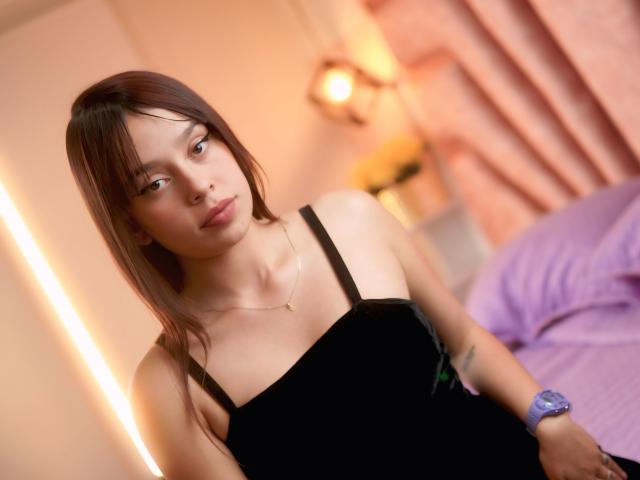 SophiaBianca with brown hair, wearing a black dress with black straps, looking at the camera, gold necklace, purple wristband, soft lighting, slight smile, bedroom setting.