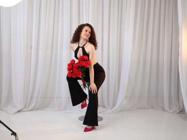 KaraCriegs with curly brown hair, black halter top, and black pants, holding red roses, wearing red heels, standing on a stool, smiling, in a studio with white curtains.