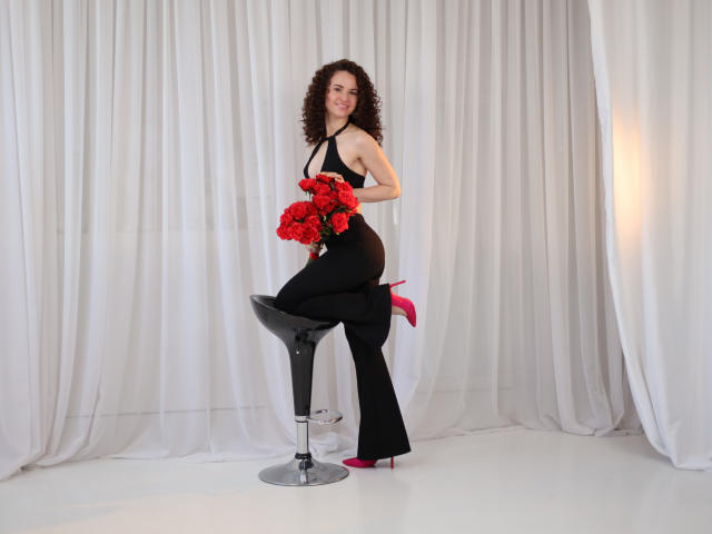 KaraCriegs, con i capelli castano ricci, un vestito nero a halter e tacchi rosa, seduta su uno sgabello nero, che tiene in mano delle rose rosse e sorride alla macchina fotografica..