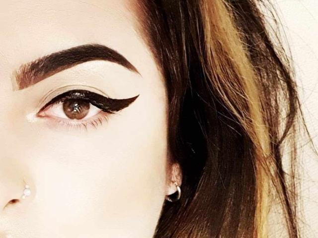 SmokingGoddess's eye with thick, dark eyebrows, brown eyelash wing, brown eye, pale skin, nose piercing with a small gem, and long brown hair with blonde highlights.