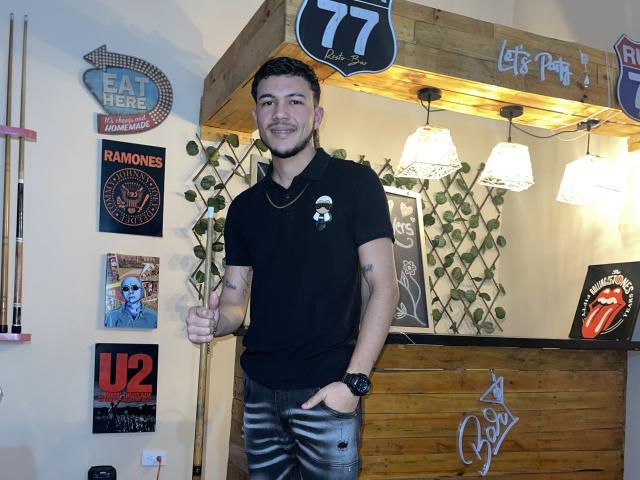 LeonardAsaga, wearing a black polo shirt and gray jeans, stands with a thumbs-up, tattoo on arm, wooden sign above, hanging lamps, and rock posters in the background.
