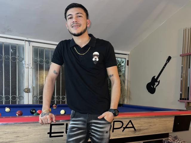 LeonardAsaga, standing by a pool table, wearing a black polo shirt with a skull emblem, black jeans, and a watch, smiling with tattoos on both arms.