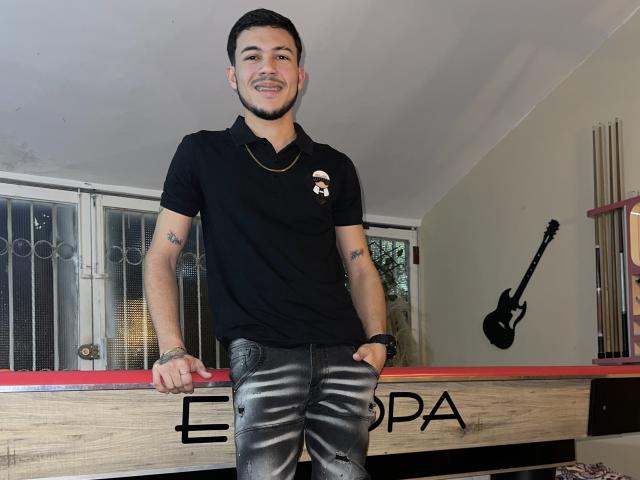 LeonardAsaga, standing, black polo shirt, gold necklace, tattoos on arms, black leather pants, short dark hair, smiling, leaning on wooden table, black guitar on wall.