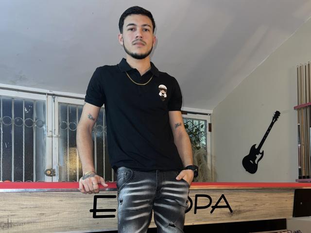 LeonardAsaga with short black hair and beard, wearing a black polo shirt and ripped black jeans, standing by a pool table with a guitar on the wall.