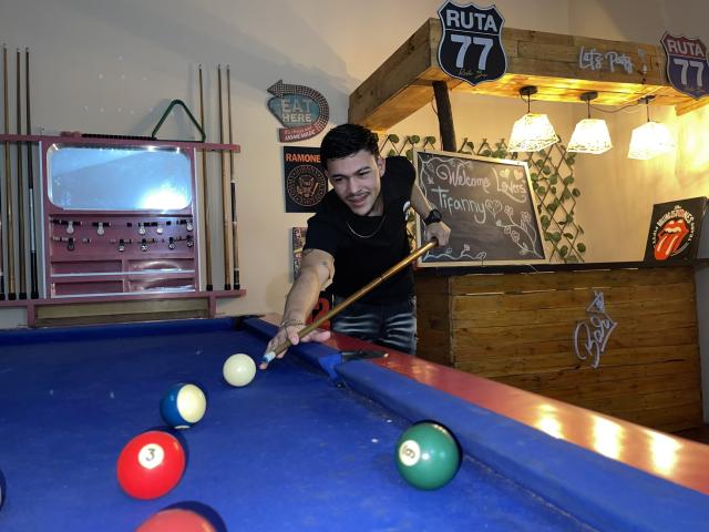 LeonardAsaga with tattoos, black shirt, and short dark hair, leans over blue pool table, aiming cue stick at white ball, surrounded by colorful pool balls and wooden bar.