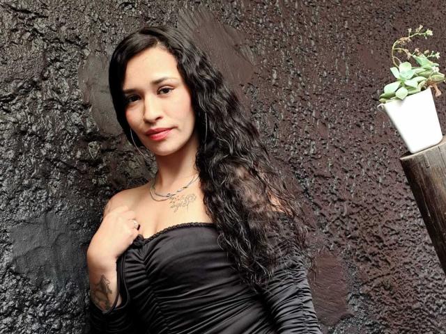 Zoey Moretti, com longos cabelos pretos e cacheados, vestindo um vestido preto com um ombro descoberto, tocando o peito, com uma pequena tatuagem no lado direito, em pé ao lado de uma planta em um vaso em uma prateleira escura..