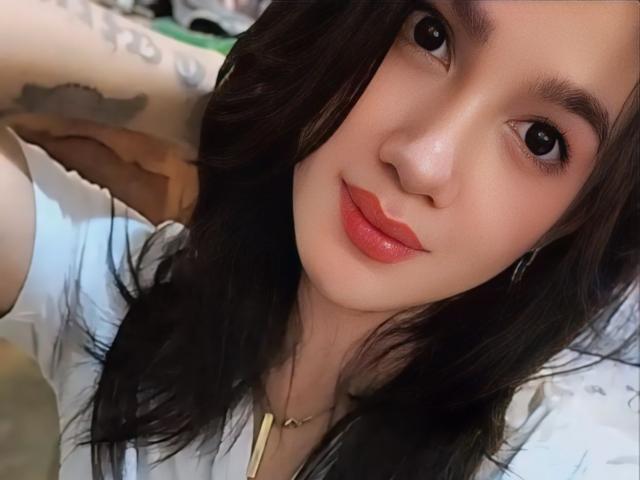 SweetNSexyD has dark hair, red lips, and a small nose. She's wearing a white shirt and a gold necklace, with tattoos on her arm. Her eyes are brown.