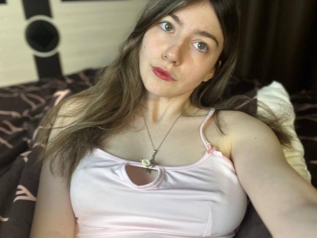 VinnaSkyHott mit einem blassrosa Satin-Oberteil, das kleine Brüste zeigt, mit langen braunen Haaren, grünen Augen, rotem Lippenstift und einer Halskette mit einer Blume, liegt auf einem Bett..