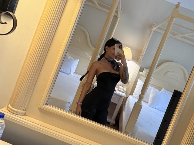 ThaliaAres trägt ein schwarzes, trägerloses Kleid, hat kleine Brüste, ein Tattoo am rechten Arm und macht ein Selfie vor dem Spiegel im Schlafzimmer. Im Hintergrund steht ein weißes Himmelbett. Sie hat schwarzes Haar zu einem Pferdeschwanz gebunden und trägt ein pinkes Armband..