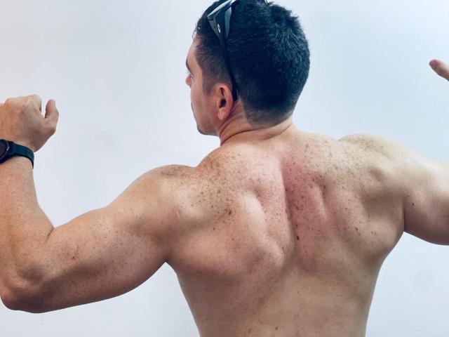 Se muestra la espalda musculosa de Jago, sin camisa, con los brazos levantados, llevando gafas de sol en la cabeza y un reloj negro en la muñeca. Su piel está salpicada de pecas y ligeramente enrojecida..
