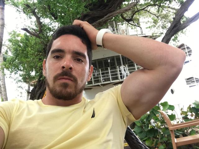 Jago, a muscular dude with dark hair and a beard, wears a yellow shirt, white wristband, and gold earrings, scratching his head, with a serious expression, in a tree-lined area.