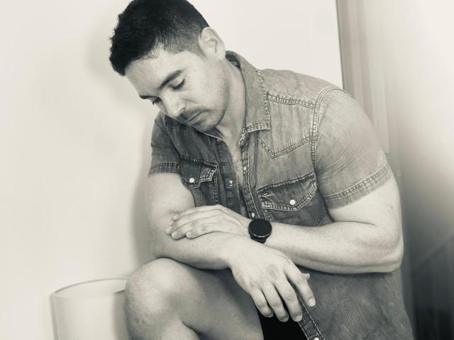 Jago, un homme corpulent avec des cheveux courts et foncés, est assis les bras croisés, portant une chemise en jean et une montre noire, et regarde vers le bas avec une expression sérieuse. Monochrome..