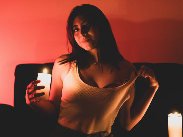 SpencerLe, con i capelli scuri, tiene una candela nella mano sinistra, tirando giù una canotta beige per mostrare i suoi seni, illuminata dalla luce rossa delle candele, seduta su un divano nero..