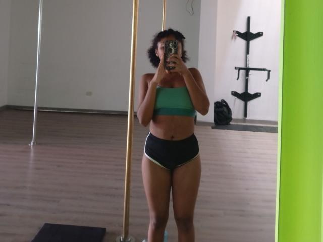 MeganWlliams with curly dark hair, wearing a green sports bra and black underwear, standing in front of a mirror, holding a phone, showing her tits and ass.
