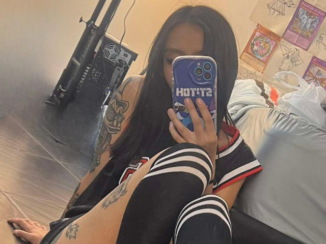 EviieGothel with long black hair, tattoos, and a phone case with "Honey" in blue, takes a selfie, showing her legs in black and white striped socks.