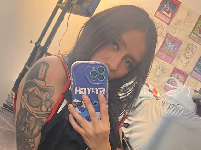 EviieGothel with black hair, holding a blue phone, showing a tattoo of a grinning character on her arm, looking at the camera, in a bedroom.