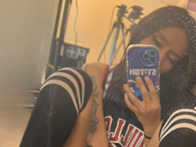 EviieGothel taking a mirror selfie, showing her tattooed thigh and wearing black and white striped knee socks, with her phone covering her face. Her phone has a blue Haribo case.