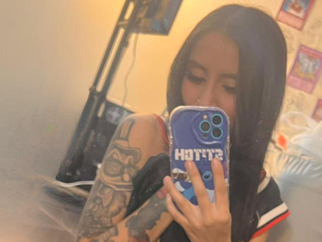 EviieGothel with long black hair, tattoos on her right arm, holding a blue phone with "Hot" on it, taking a mirror selfie, showing her red bra strap.