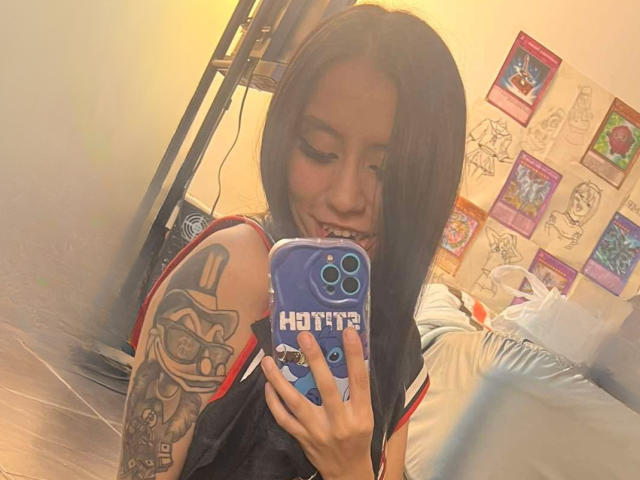 EviieGothel with long black hair, smiling, holding a blue phone with "Hot" case, showing a tattoo on her right arm, in a mirror selfie. Her tits are slightly visible.
