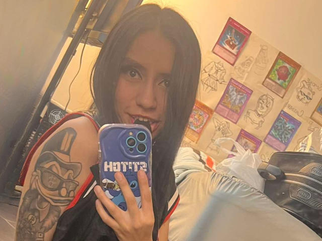 EviieGothel with long dark hair, tongue out, holding a blue iPhone, showing a tattoo on her right arm. She's wearing a black and red top, looking at the camera.