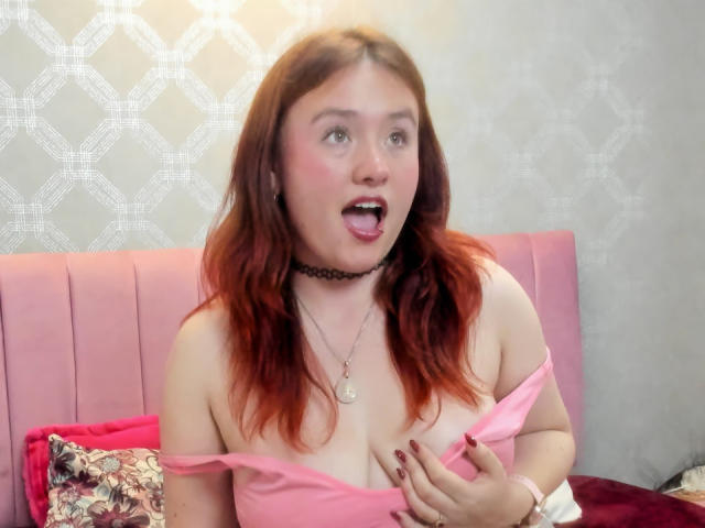 AliaMillers with red hair, choker, and pink top, exposing her tits, mouth open, looking surprised, hand on her chest, nail polish, and a pink bed behind her.