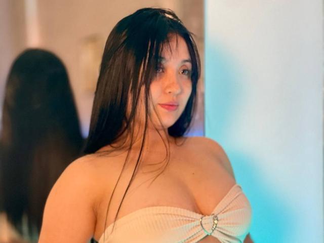 LunaBellaHotty with long dark hair, pale skin, and big tits in a beige strapless top, looking at the camera with a slight smile. Mirror reflection in background.
