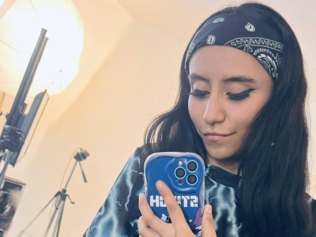 EviieGothel with long black hair, black bandana, and blue tie-dye shirt, looking down at her blue iPhone, has sharp winged eyeliner and a slight smirk.