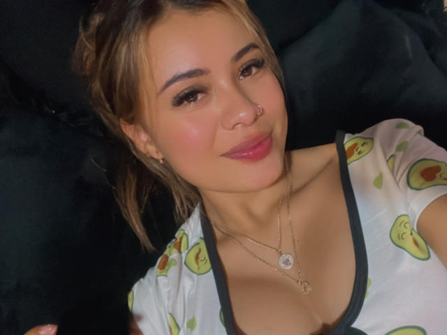 ArianaBellee with brown hair, nose ring, and pink lipstick, wearing a white shirt with avocado prints, showing her tits, and two necklaces, smiling at the camera.