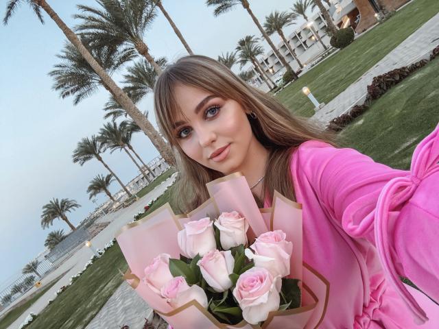 DreamySamantha with pink roses, holding bouquet in pink jacket, palm trees, grass, and pathway in background, selfie, blue eyes, long brown hair, pink jacket tied at wrist.