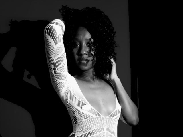 Black-and-white photo of ChocolateSipp with curly hair, wearing a white, see-through, long-sleeve top, exposing small tits, hand in hair, shadow on wall, serious expression.