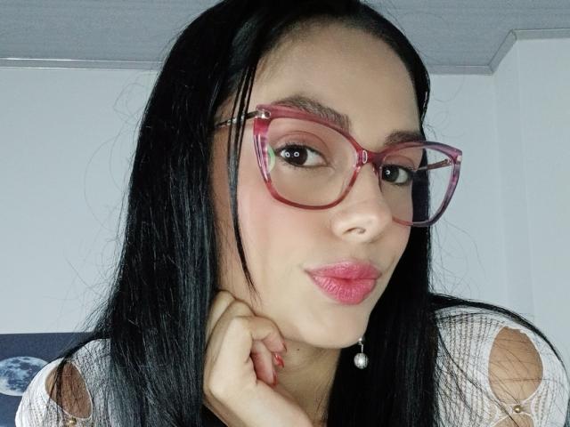 AnaisHilton with red glasses, black hair, pink lipstick, and a pearl earring, pouting her lips with her hand under her chin. She's wearing a white top with cut-out sleeves.