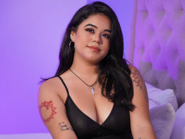 Victoria Boston, com cabelo preto, pele média, tatuagens nos braços, vestindo um vestido preto, mostrando os seios, brincos grandes, e um colar, sorrindo em um quarto iluminado por luzes roxas..