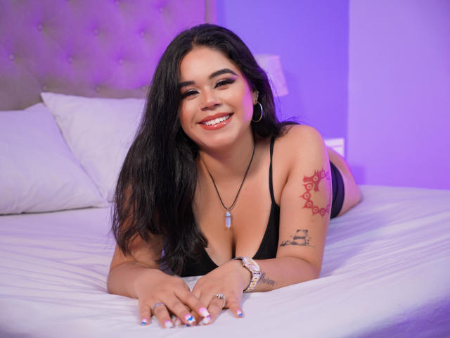 Victoria Boston, con cabello largo y negro, sonriendo, vestida con lencería negra, acostada en una cama, mostrando tatuajes en su brazo y llevando un collar con una piedra azul..