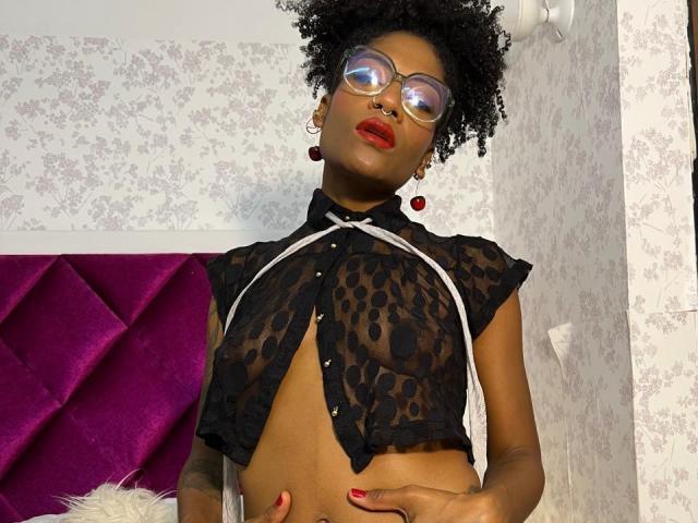 Eimy Hadid, avec des cheveux bouclés, des lunettes, du rouge à lèvres rouge et un haut noir transparent à motifs léopard, montrant de petites seins, le ventre et des boucles d'oreilles rouges, se tient dans une pièce avec du papier peint floral blanc..