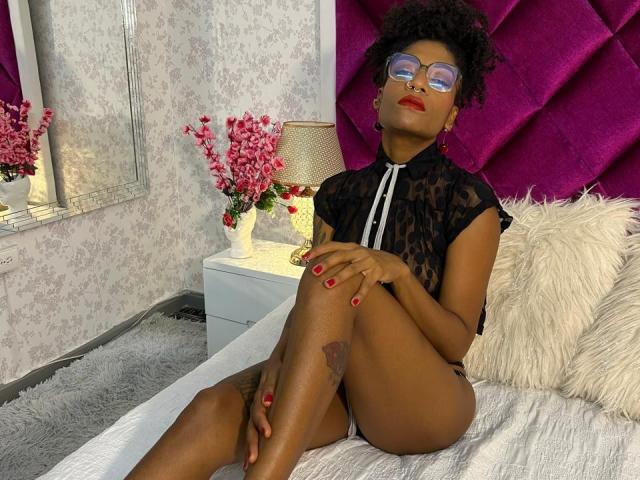 Eimy Hadid, con piel oscura, lápiz labial rojo y gafas, vestida con una blusa negra de encaje, sentada en una cama con cabecera morada, con las piernas cruzadas, esmalte de uñas rojo y un cojín blanco..