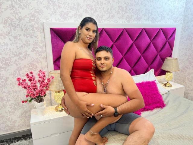 VioletandMaxC with tan skin and brown hair, wearing a red cut-out dress, sits on MaxC's lap, who is shirtless and in gray shorts, in a bedroom.