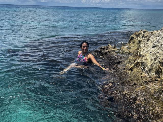 HeraDiosa with dark hair, wearing a colorful one-piece, is in the clear blue ocean, sitting on rocky shore, smiling, with her ass and legs in the water.