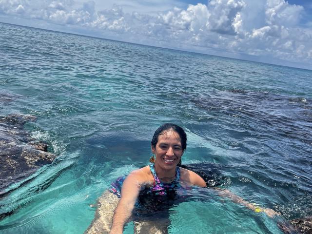 HeraDiosa with dark hair, smiling, wearing a colorful bikini, wading in clear blue ocean water, with rocky underwater patches, and a cloudy sky above. Her tits are visible.