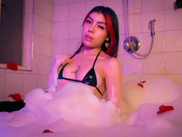 SamantaDeVous in een bubbelbad, gekleed in een zwart micro-bikini, waarbij haar borsten zichtbaar zijn, met rode rozenblaadjes en paars licht. Ze houdt een koord van haar bikini-top vast..
