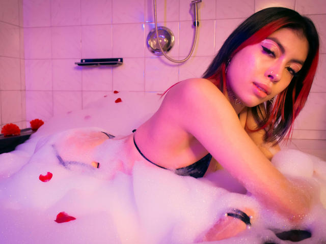 SamantaDeVous dans un bain moussant, en lingerie noire, avec des cheveux noirs et rouges, un éclairage rose et violet, des pétales de rose, allongée sur le côté, regardant la caméra, seins visibles, expression séduisante..