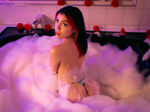 SamantaDeVous en una bañera, mirando hacia atrás con reflejos rojos en su cabello oscuro, mostrando su trasero y espalda, usando una tanga negra, con iluminación morada y rosa..