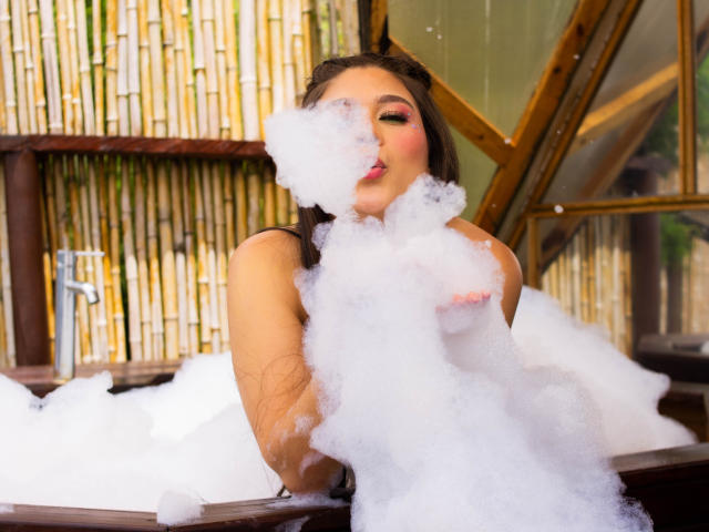 LindaJimenex, with soapy bubbles covering her tits and mouth, sits in a wooden bathtub, her dark hair wet, eyes partially closed, in a bamboo-walled room.