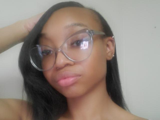 NadiaaSexy with dark brown skin, black straight hair, clear glasses, pink lipstick, and a serious expression, raising her right hand to her head.