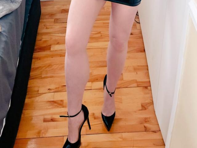 TitanicTits' legs, wearing black high heels with ankle straps, standing on a wooden floor. She's got pale skin, and her short black dress is just above her ass.