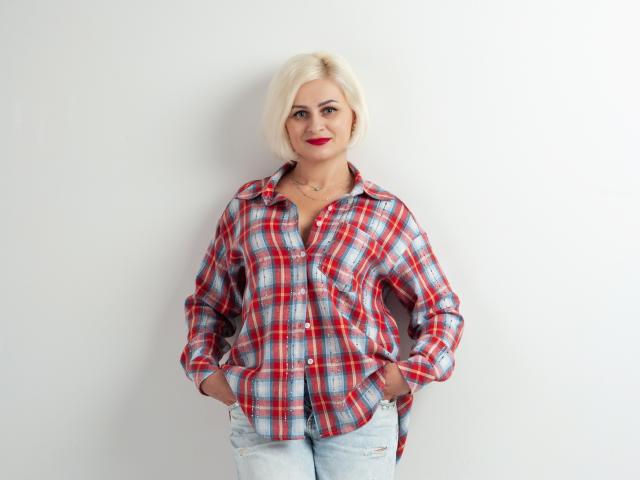OliviaHorst with blonde bob, red lipstick, wearing red plaid shirt, hands in jeans pockets, standing against white wall, smiling confidently.